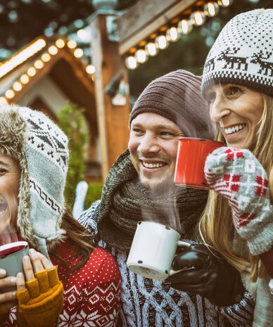 Three friends wearing knit hats sharing hot drinks at a Christmas market.