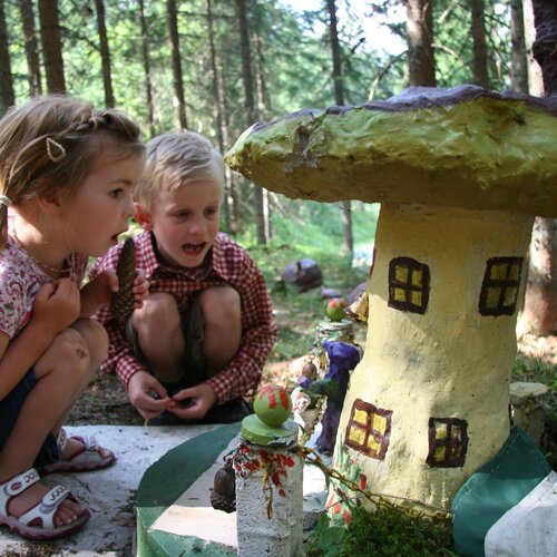 Two children crouching and inspecting a painted mushroom house in a forest