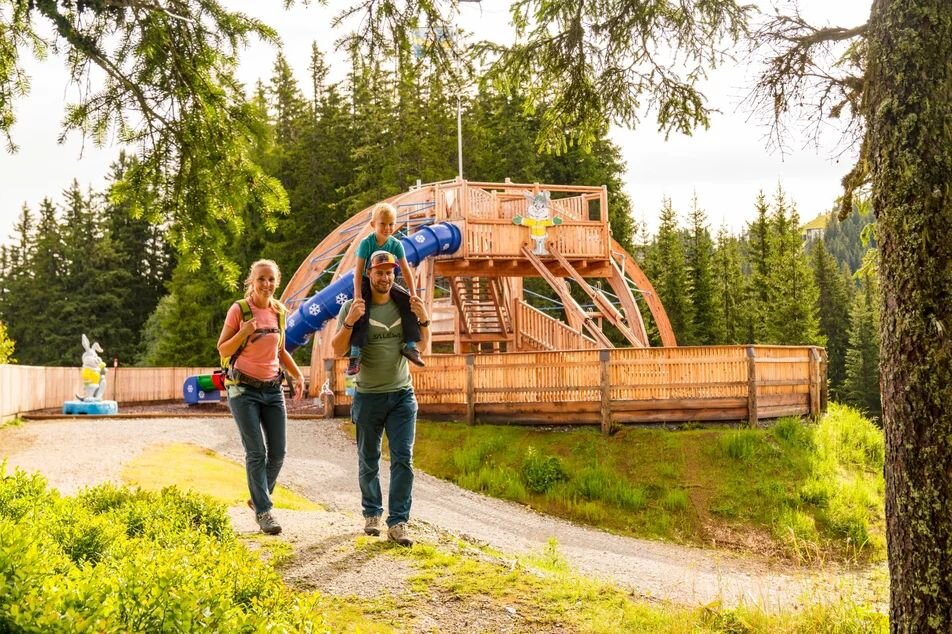 Family with child on shoulders walking beside wooden playground