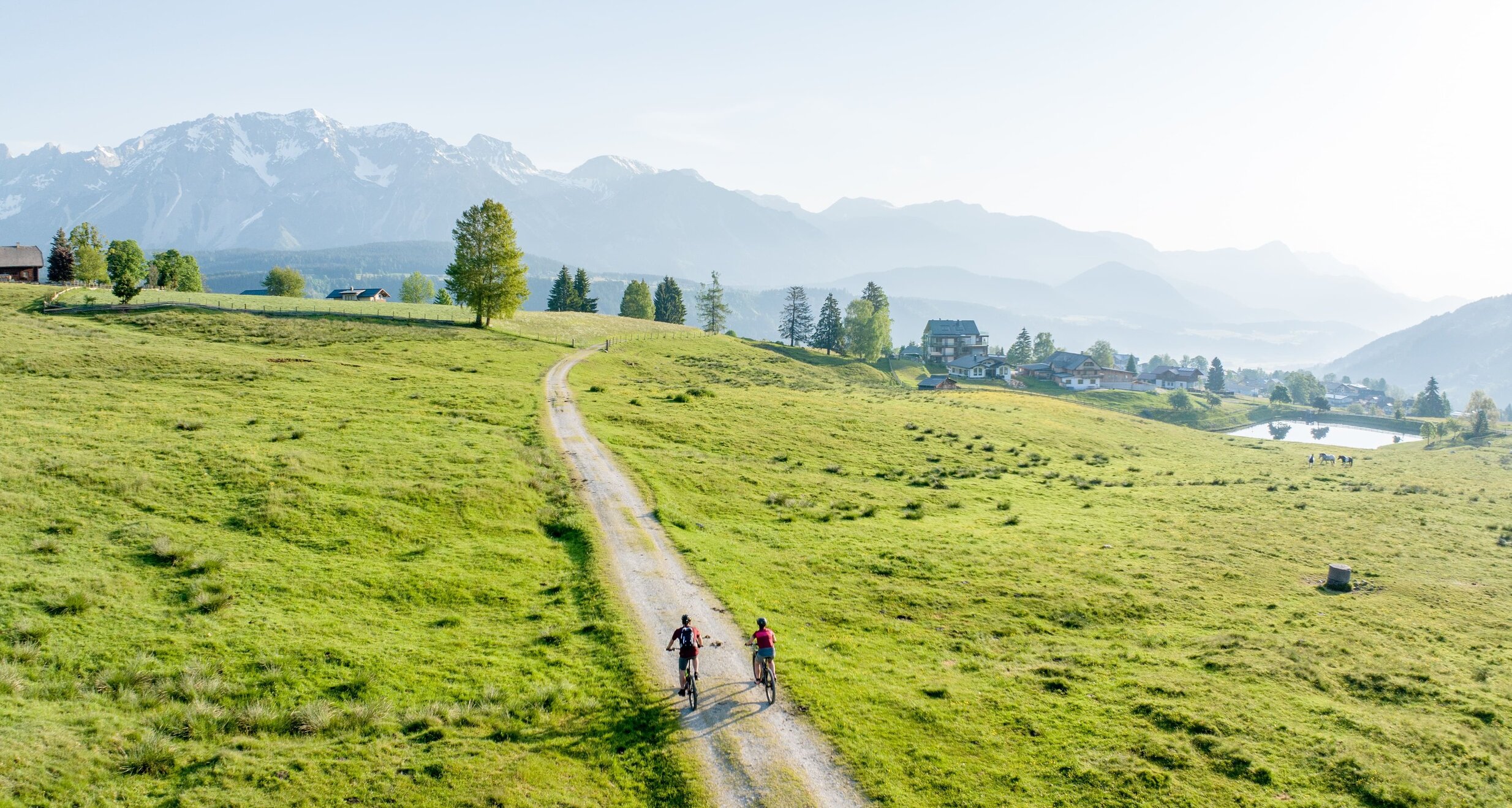 Two cyclists on a dirt path through green pastures with distant mountains