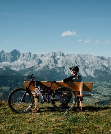 Cyclist wearing helmet sits on bench overlooking mountains with bike.