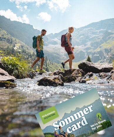 Two hikers cross a rocky stream in alpine valley; Sommer Card visible.