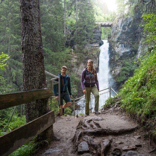Two hikers on a forest trail beside a waterfall.