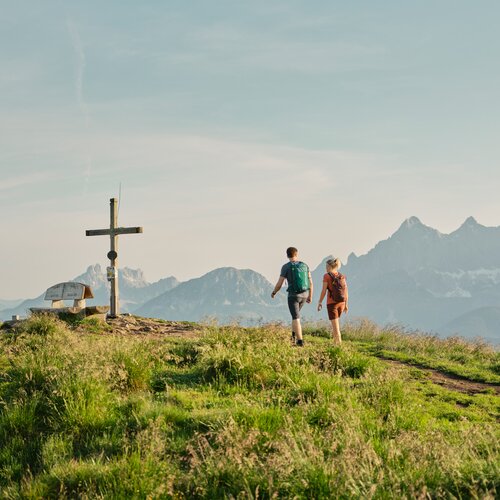 Hikers with backpacks walk along grassy summit trail near a wooden cross. | © Peter Burgstsaller