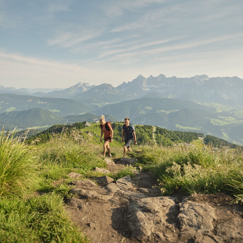 Two hikers ascend a rocky mountain trail with valley and peaks