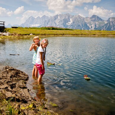 Two girls stand in a shallow alpine lake by a grassy bank.