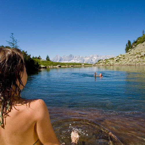 Person in green bikini sits on rocky lakeshore watching swimmers