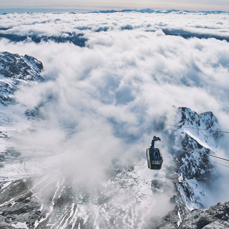 Cable car suspended above snow-covered mountains and clouds. | © Robert Maybach