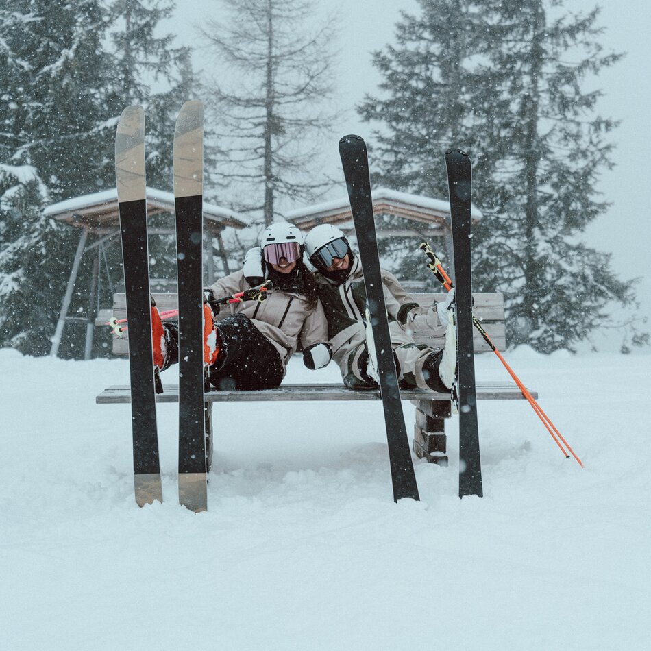 Two skiers sitting on a bench in the snow with skis nearby.