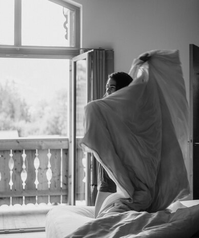 Person lifting blanket on bed beside open balcony door