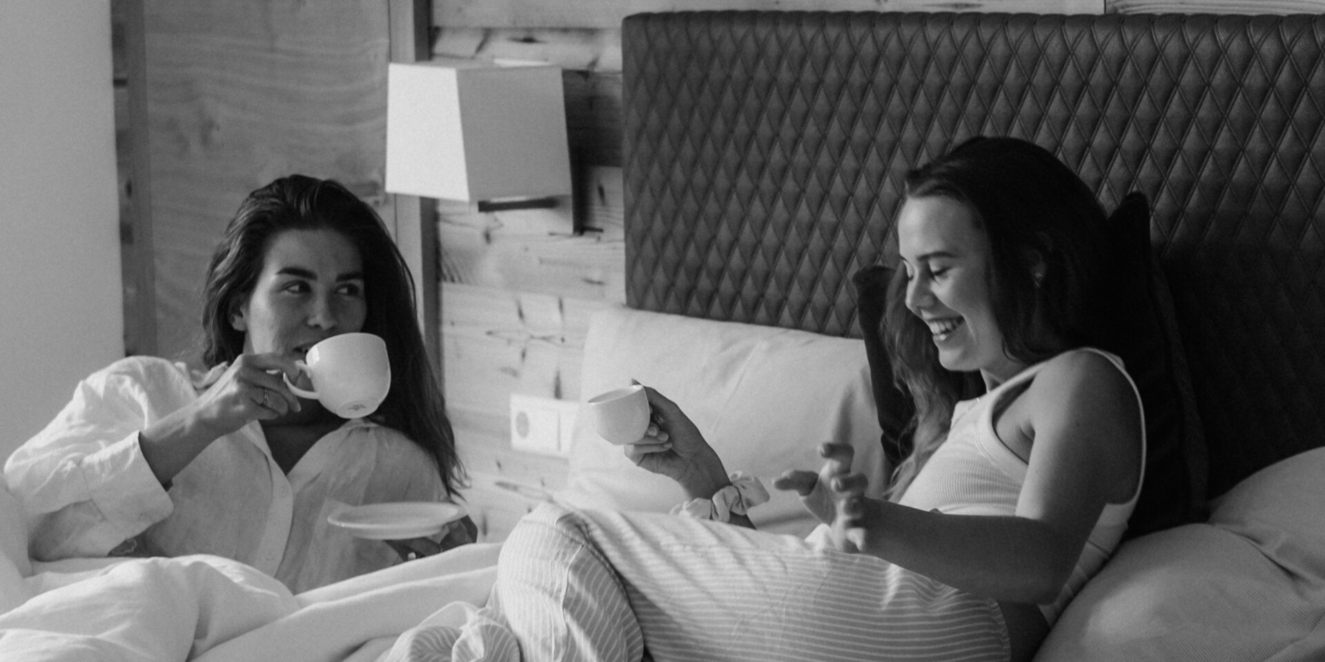 Two women in bed sharing coffee and smiling.