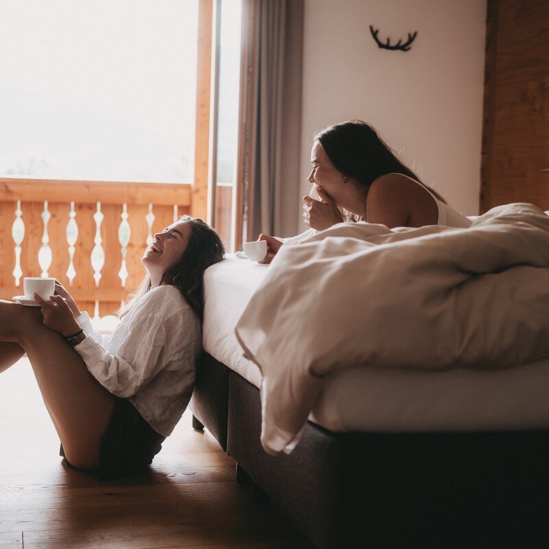 Two women laughing with coffee cups near the bed