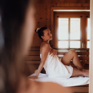 Woman in white towel sits on sauna bench by window.