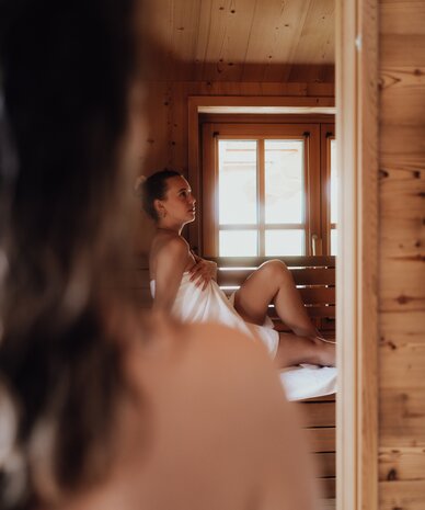 Woman in towel sits on wooden sauna bench beside window.