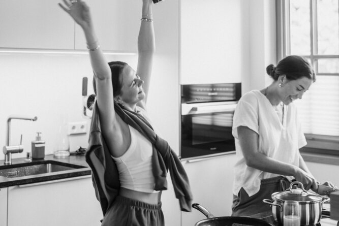 Two women in a bright modern kitchen; one raises arms.