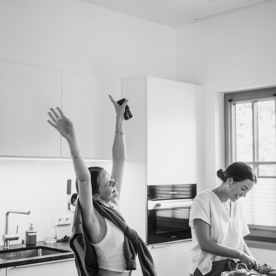 Two women in a bright modern kitchen; one raises arms.