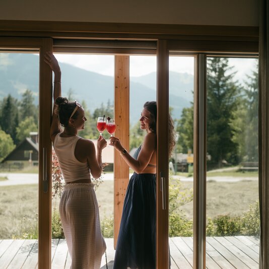 Two women toast red drinks on a wooden deck