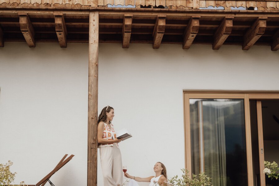 Two women on a wooden house patio, reading and toasting