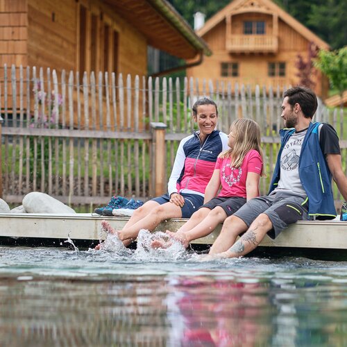 Family of three sitting on a dock, feet splashing water | © RAPHAELGABAUER.COM