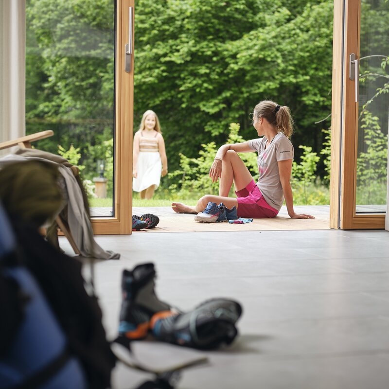 Woman seated on indoor floor by open doors, watching outside. | © RAPHAELGABAUER.COM
