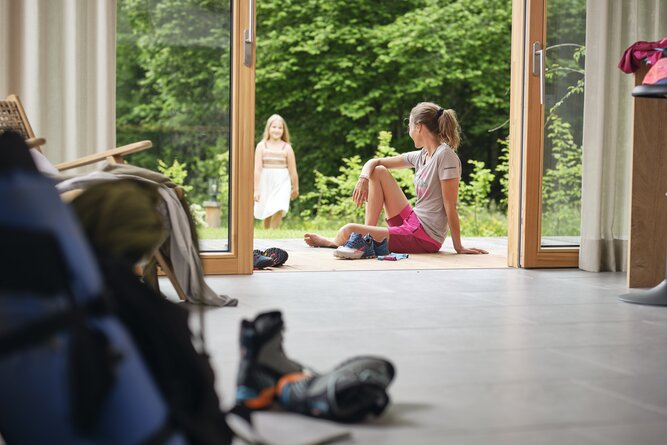 Woman seated on indoor floor by open doors, watching outside. | © RAPHAELGABAUER.COM