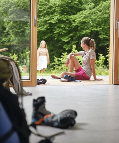 Woman seated on indoor floor by open doors, watching outside. | © RAPHAELGABAUER.COM