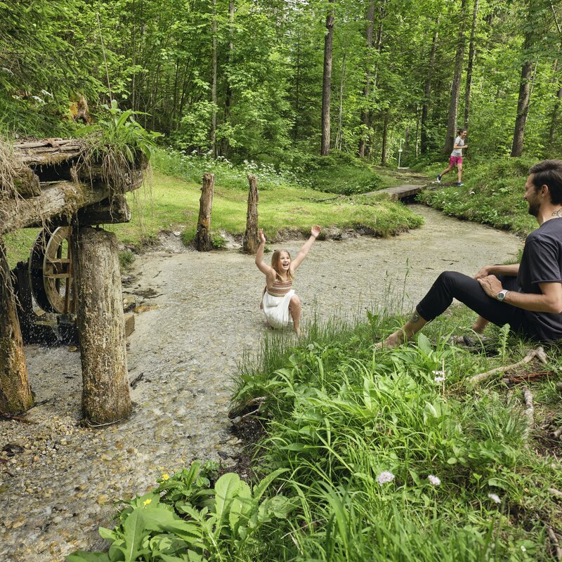 Girl splashes in shallow stream while man sits nearby. | © RAPHAELGABAUER.COM